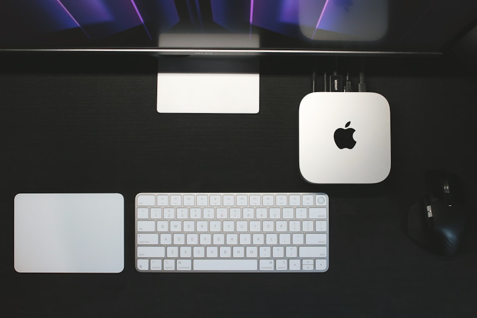 A Mac mini on a dark desk between a keyboard and a trackpad, lit from above by the glow of the monitor it's plugged into.